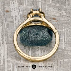The "Diana" ring in 18K yellow gold with a 1.08-carat Montana sapphire is displayed on a metallic, patterned background featuring the "Earth's Treasury" logo at the bottom.