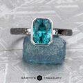 The Stackable "Emma" ring in hammered 14K white gold, featuring a 1.37-carat teal tourmaline, is shown upright on textured blue stone. The Earth's Treasury logo appears at the bottom.