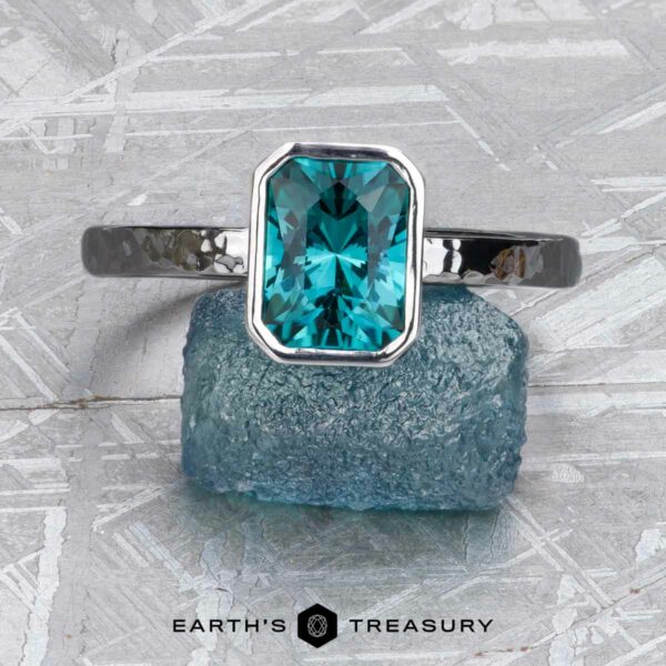 The Stackable "Emma" ring in 14K white gold, hammered, with 1.37-carat tourmaline The Stackable "Emma" ring in hammered 14K white gold, featuring a 1.37-carat teal tourmaline, is shown upright on textured blue stone. The Earth's Treasury logo appears at the bottom.