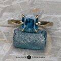 A 14K yellow gold "Nelisa" ring with a 2.36-carat Montana sapphire rests on a rough blue crystal atop a textured gray surface. The text "EARTH'S TREASURY" appears at the bottom.