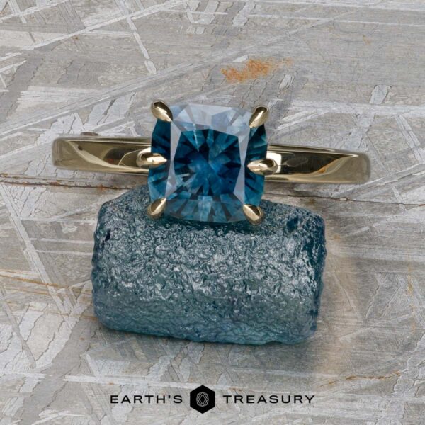The "Nelisa" ring in 14K yellow gold with 2.36-carat Montana sapphire A 14K yellow gold "Nelisa" ring with a 2.36-carat Montana sapphire rests on a rough blue crystal atop a textured gray surface. The text "EARTH'S TREASURY" appears at the bottom.
