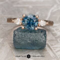The "Clematis" ring in 14K rose gold, featuring a 1.71-carat round Montana sapphire and two pear-shaped side stones, is displayed on rough blue stone with "Earth's Treasury" below.