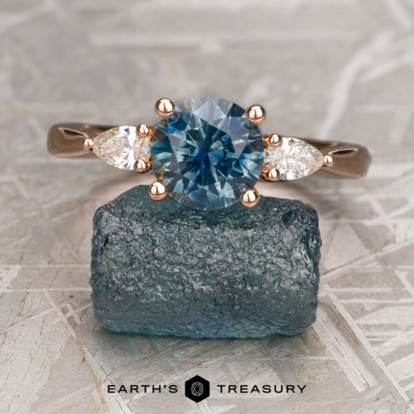 The "Clematis" in 14K rose gold with 1.71-carat Montana sapphire The "Clematis" ring in 14K rose gold, featuring a 1.71-carat round Montana sapphire and two pear-shaped side stones, is displayed on rough blue stone with "Earth's Treasury" below.