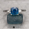 The "Erythia" ring in platinum, featuring a 1.89-carat blue Montana sapphire and two side diamonds, is displayed on a blue mineral specimen with a gray background and the Earth's Treasury logo at the bottom.