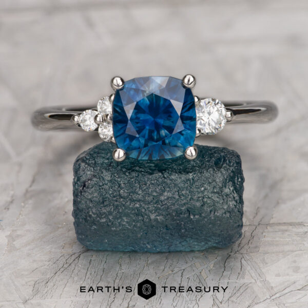 The "Erythia" ring in platinum with 1.89-carat Montana sapphire The "Erythia" ring in platinum, featuring a 1.89-carat blue Montana sapphire and two side diamonds, is displayed on a blue mineral specimen with a gray background and the Earth's Treasury logo at the bottom.