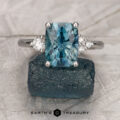 The "Erythia" ring in 14K white gold features a 3.29-carat Montana sapphire framed by sparkling diamonds, resting on a rough blue stone. The text "EARTH'S TREASURY" appears below, capturing the enchanting allure of Erythia.