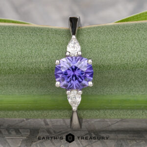 The "Clematis" Ring in platinum features a 1.62-carat round Madagascar sapphire at the center, accented by two pear-shaped clear stones, shown displayed on a green leaf.