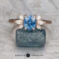 The "Liriope" ring in 14k rose gold with a 1.40-carat Montana sapphire and marquise-cut diamond accents is displayed on a rough blue stone against a textured gray background. "EARTH'S TREASURY" appears below.