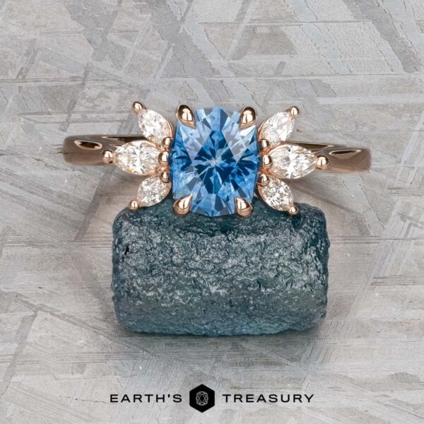 The "Liriope" ring in 14k rose gold with 1.40-carat Montana sapphire The "Liriope" ring in 14k rose gold with a 1.40-carat Montana sapphire and marquise-cut diamond accents is displayed on a rough blue stone against a textured gray background. "EARTH'S TREASURY" appears below.