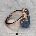 The "Liriope" ring, crafted in 14k rose gold and set with a 1.40-carat Montana sapphire, is showcased atop a rough dark mineral against a textured gray background.
