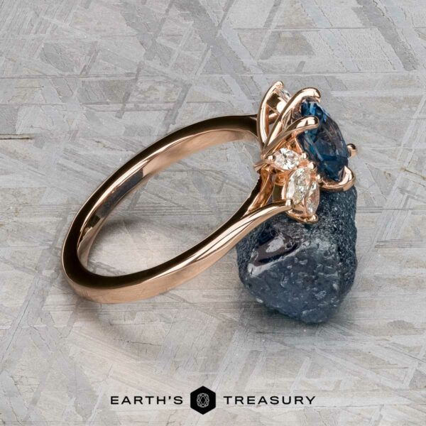 The "Liriope" ring in 14k rose gold with 1.40-carat Montana sapphire The "Liriope" ring, crafted in 14k rose gold and set with a 1.40-carat Montana sapphire, is showcased atop a rough dark mineral against a textured gray background.