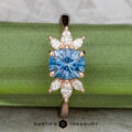 Showcasing the "Liriope" ring in 14k rose gold, this piece features a 1.40-carat Montana sapphire at its center with marquise and pear-shaped clear stones, elegantly displayed on a green background.