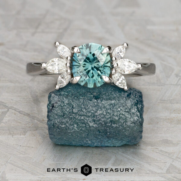 The "Liriope" ring in 14k white gold with 1.23-carat Montana sapphire The "Liriope" ring in 14k white gold with a 1.23-carat Montana sapphire and four marquise-cut clear stones rests on rough blue mineral. "EARTH'S TREASURY" logo appears below, showcasing unique rings inspired by nature's beauty.
