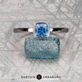 The McMinn, Richard and Gomez, Eric stackable silver ring, featuring a cushion-cut blue gemstone, rests on a raw blue crystal atop a patterned metallic surface with "Earth's Treasury" below.