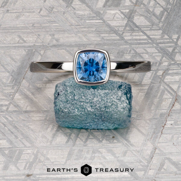 McMinn, Richard and Gomez, Eric The McMinn, Richard and Gomez, Eric stackable silver ring, featuring a cushion-cut blue gemstone, rests on a raw blue crystal atop a patterned metallic surface with "Earth's Treasury" below.