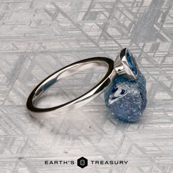 McMinn, Richard and Gomez, Eric A McMinn, Richard and Gomez, Eric solitaire ring with an oval blue gemstone sits on a rough blue stone atop a geometric surface with the "Earth's Treasury" logo—ideal as a stackable or elegant solitaire ring.