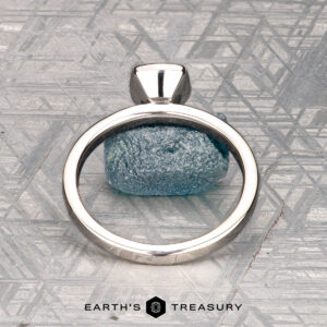 A solitaire ring with a rough blue gemstone is shown on a gray patterned surface, labeled "McMinn, Richard and Gomez, Eric" below.