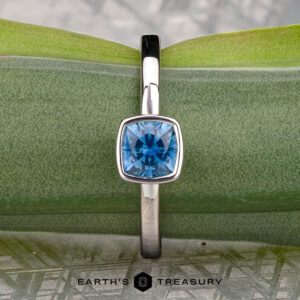 A stackable silver ring with a square bezel-set blue gemstone is displayed on a green leaf, with "McMinn, Richard and Gomez, Eric" written below.