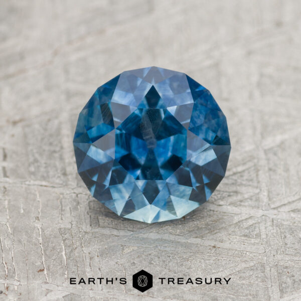 1.78-Carat Medium Blue Montana Sapphire (Heated) A 1.78-carat medium blue heated Montana Sapphire, round and faceted, rests on a textured gray surface with the "Earth's Treasury" logo and text at the bottom.