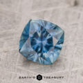 A 1.82-carat blue-green Montana sapphire (heated) is displayed on a textured surface with the "EARTH'S TREASURY" logo and text beneath it.