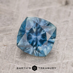 A 1.82-carat blue-green Montana sapphire (heated) is displayed on a textured surface with the "EARTH'S TREASURY" logo and text beneath it.