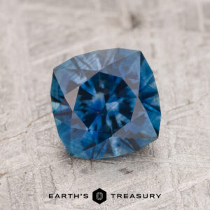 A faceted, square-cut 1.81-Carat Deep Blue Montana Sapphire (Heated) is displayed on a textured gray surface, with the Earth’s Treasury logo beneath.