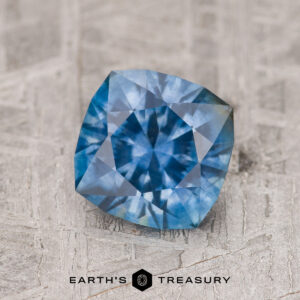 A 1.54-carat medium blue Montana sapphire (heated) with a cushion cut is shown on a textured surface, featuring the "EARTH'S TREASURY" text and logo at the bottom.