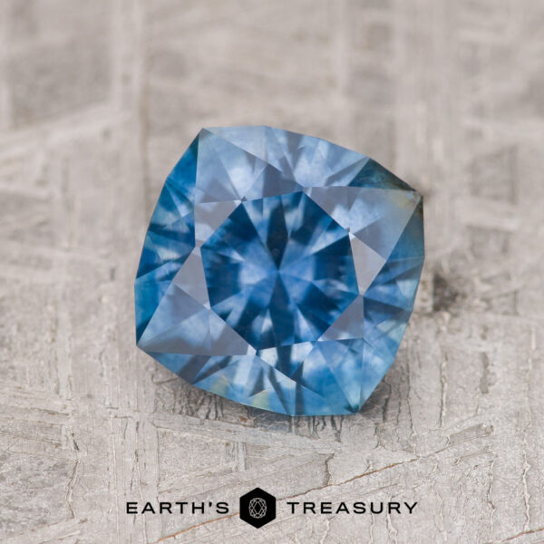 1.54-Carat Medium Blue Montana Sapphire (Heated) A 1.54-carat medium blue Montana sapphire (heated) with a cushion cut is shown on a textured surface, featuring the "EARTH'S TREASURY" text and logo at the bottom.