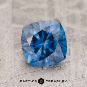 A 1.47-carat medium blue heated Montana Sapphire is displayed on a textured gray surface featuring the "Earth's Treasury" logo at the bottom.