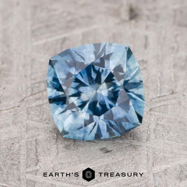 1.53-Carat Aqua Blue Montana Sapphire (Heated) A 1.53-carat heated Aqua Blue Montana Sapphire with a cushion cut and intricate facets rests on a textured gray surface. "EARTH'S TREASURY" and its logo appear at the bottom of the image.
