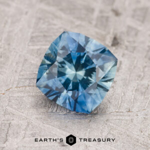 A 1.35-carat teal blue Montana sapphire (heated), cushion-cut and beautifully faceted, rests on a textured light background above the "EARTH'S TREASURY" logo.