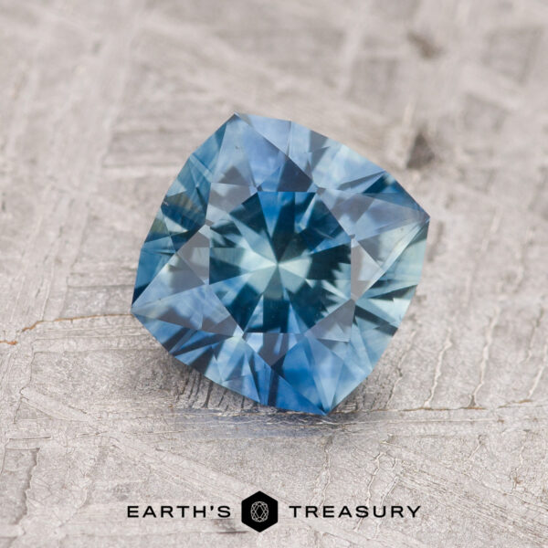 1.35-Carat Teal Blue Montana Sapphire (Heated) A 1.35-carat teal blue Montana sapphire (heated), cushion-cut and beautifully faceted, rests on a textured light background above the "EARTH'S TREASURY" logo.
