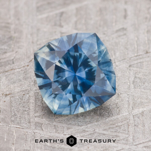 1.79-Carat Teal Blue Montana Sapphire (Heated) Product SNYTHR-11041-1200-BW, a Teal Blue Montana Sapphire with a star-like internal pattern, is shown on a textured gray surface with the "Earth's Treasury" logo beneath.