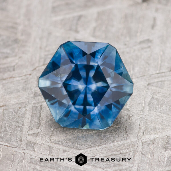 A 1.46-carat deep blue Montana sapphire (heated), faceted in an octagonal shape, rests on a textured gray surface with the Earth’s Treasury logo below.