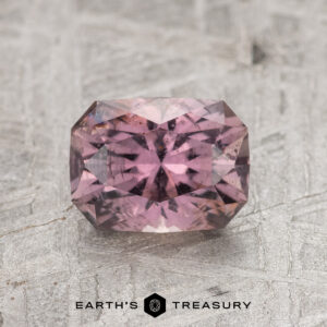 A faceted, rectangular 1.45-Carat Mauve Purple Montana Sapphire with internal reflections rests on a textured gray surface, with the "Earth's Treasury" logo and text below.