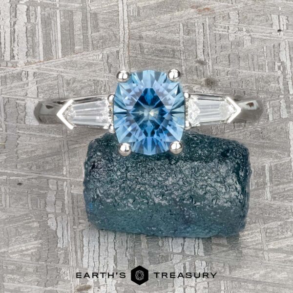 The "Mandevilla" ring in platinum with 1.61-carat Montana sapphire The "Mandevilla" ring in platinum features a 1.61-carat Montana sapphire cushion-cut center stone and two baguette diamonds, displayed on a blue mineral with the Earth's Treasury logo below.