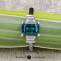 A rectangular blue "Cyanea" three-stone ring in 18K white gold with a 2.69-carat Montana sapphire is displayed on a green plant stem, the Earth’s Treasury logo shown below.