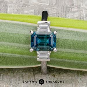 A rectangular blue "Cyanea" three-stone ring in 18K white gold with a 2.69-carat Montana sapphire is displayed on a green plant stem, the Earth’s Treasury logo shown below.