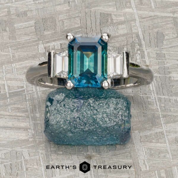 The "Cyanea" in 18K white gold with 2.69-Carat Montana Sapphire The "Cyanea" ring in 18K white gold features a 2.69-carat Montana Sapphire flanked by two clear stones, displayed on a rough blue crystal with the Earth's Treasury logo below.