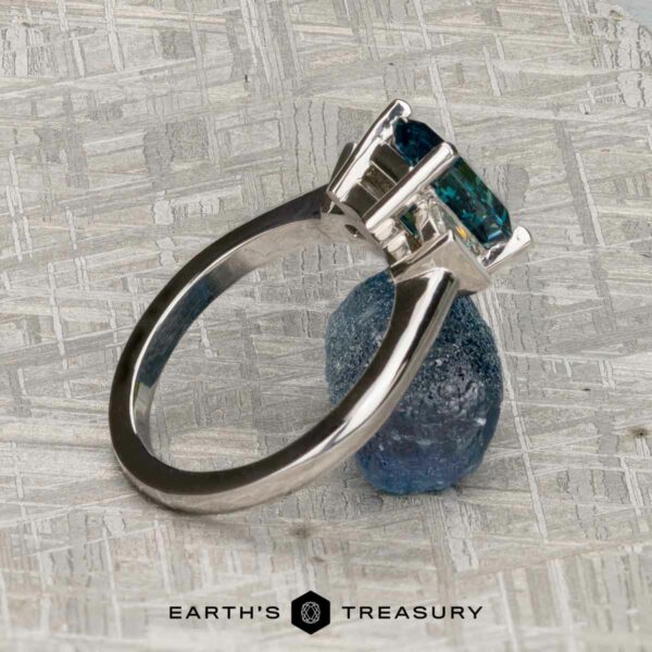 The "Cyanea" in 18K white gold with 2.69-Carat Montana Sapphire The "Cyanea" ring in 18K white gold, featuring a 2.69-carat rectangular Montana sapphire, stands upright on a textured blue stone atop a gray patterned surface, radiating modern elegance.