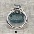 The "Cyanea" three-stone ring in 18K white gold showcases a 2.69-carat Montana sapphire and three rectangular faceted stones, photographed from above on a textured gray surface.