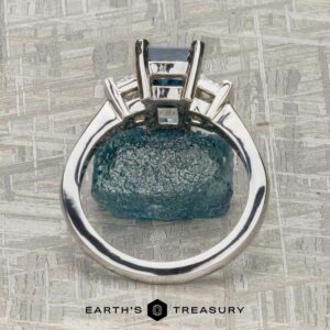 The "Cyanea" three-stone ring in 18K white gold showcases a 2.69-carat Montana sapphire and three rectangular faceted stones, photographed from above on a textured gray surface.
