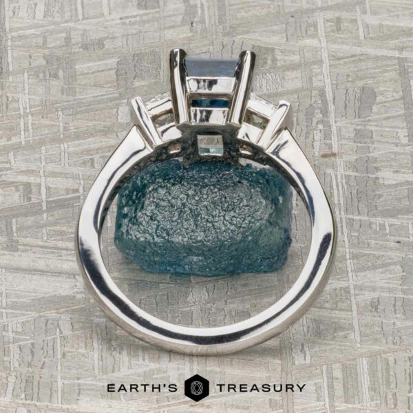 The "Cyanea" in 18K white gold with 2.69-Carat Montana Sapphire The "Cyanea" three-stone ring in 18K white gold showcases a 2.69-carat Montana sapphire and three rectangular faceted stones, photographed from above on a textured gray surface.
