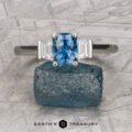 A rectangular blue "Cyanea" ring in platinum with a 1.23-carat Montana sapphire and two clear side stones rests on a rough blue mineral atop textured gray, with the "Earth's Treasury" logo visible beneath this stunning three-stone display.