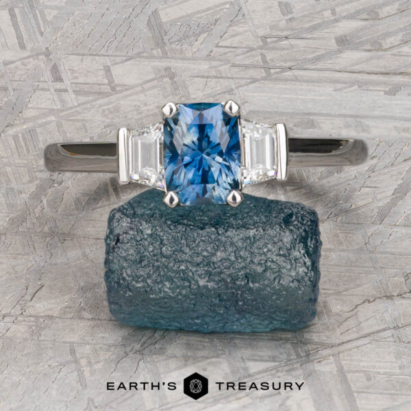 The "Cyanea" in platinum with 1.23-carat Montana sapphire A rectangular blue "Cyanea" ring in platinum with a 1.23-carat Montana sapphire and two clear side stones rests on a rough blue mineral atop textured gray, with the "Earth's Treasury" logo visible beneath this stunning three-stone display.