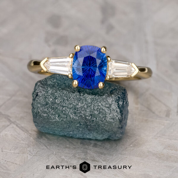 The "Mandevilla" ring in 18K yellow gold with 1.21-carat Ceylon sapphire The "Mandevilla" ring in 18K yellow gold features a 1.21-carat Ceylon sapphire center and tapered baguette diamonds, displayed on a rough blue-green stone against a textured gray background.