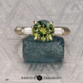 The "Mandevilla" ring in 14k yellow gold features a 1.80-carat Australian sapphire flanked by two rectangular side stones, shown on a raw crystal against a patterned surface. "Earth's Treasury" appears at the bottom.