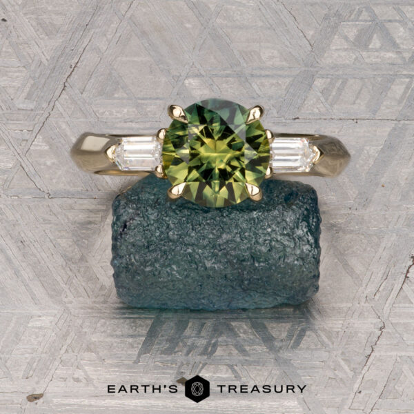 The "Mandevilla" ring in 14k yellow gold with 1.80-carat Australian sapphire The "Mandevilla" ring in 14k yellow gold features a 1.80-carat Australian sapphire flanked by two rectangular side stones, shown on a raw crystal against a patterned surface. "Earth's Treasury" appears at the bottom.
