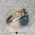 The "Mandevilla" ring, crafted in 14k yellow gold and set with a 1.80-carat Australian sapphire, sits on a rough blue stone against a patterned gray background. The "Earth's Treasury" logo appears below, inspired by Mandevilla flower elegance.