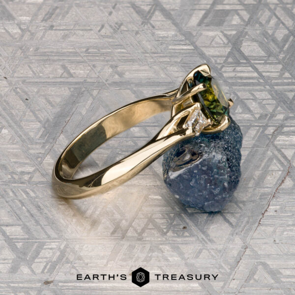 The "Mandevilla" ring in 14k yellow gold with 1.80-carat Australian sapphire The "Mandevilla" ring, crafted in 14k yellow gold and set with a 1.80-carat Australian sapphire, sits on a rough blue stone against a patterned gray background. The "Earth's Treasury" logo appears below, inspired by Mandevilla flower elegance.